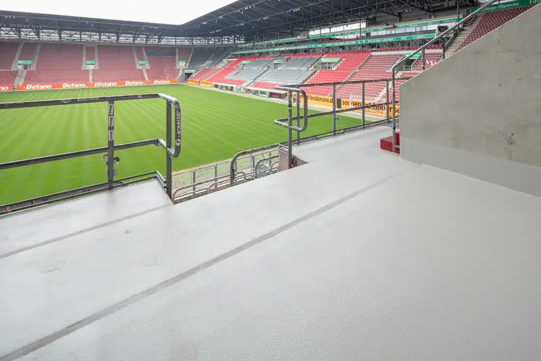 Blick von einer frisch beschichteten Tribünenplattform mit Metallgeländer auf das Spielfeld und die roten Ränge eines Fußballstadions.