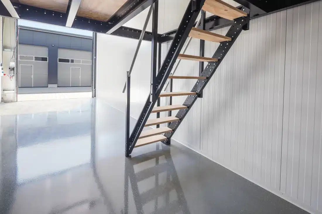 Industrietreppe aus schwarzem Stahl mit Holzstufen führt auf ein Mezzanin in einer hellen Lagerhalle mit Epoxidharzboden.