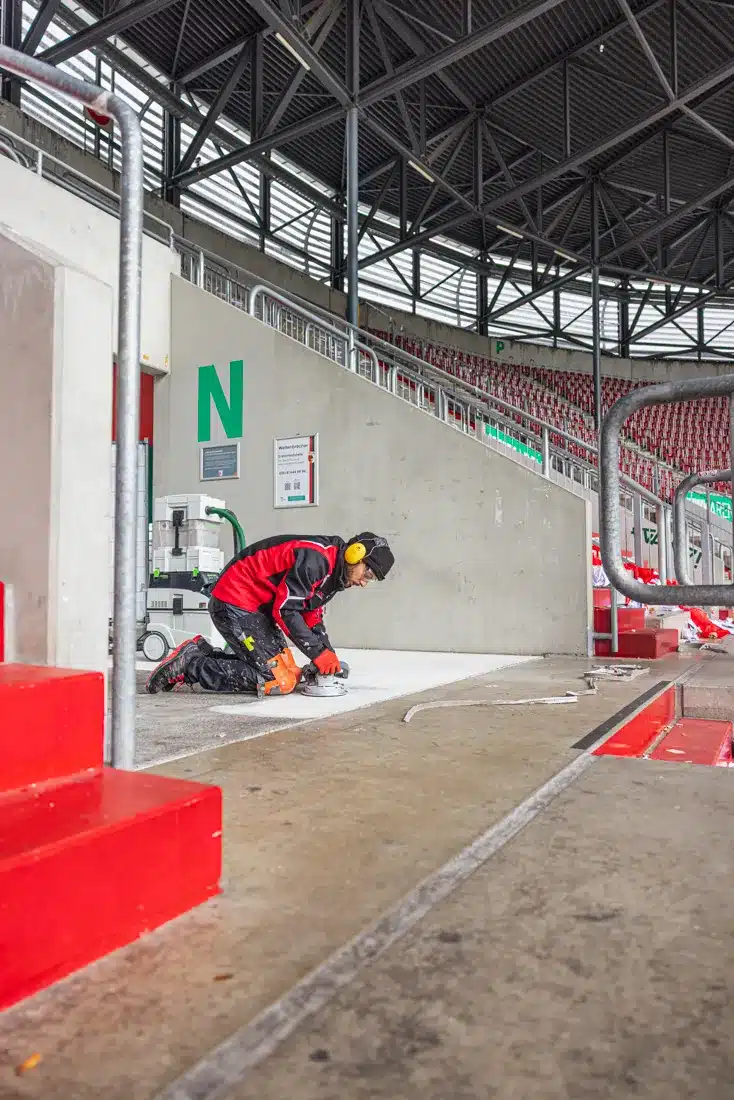 Sanierungsarbeiten im Stadion: Facharbeiter schleift kniend den Bodenbelag eines Tribünenaufgangs unter einer Stahlrohr-Dachkonstruktion.