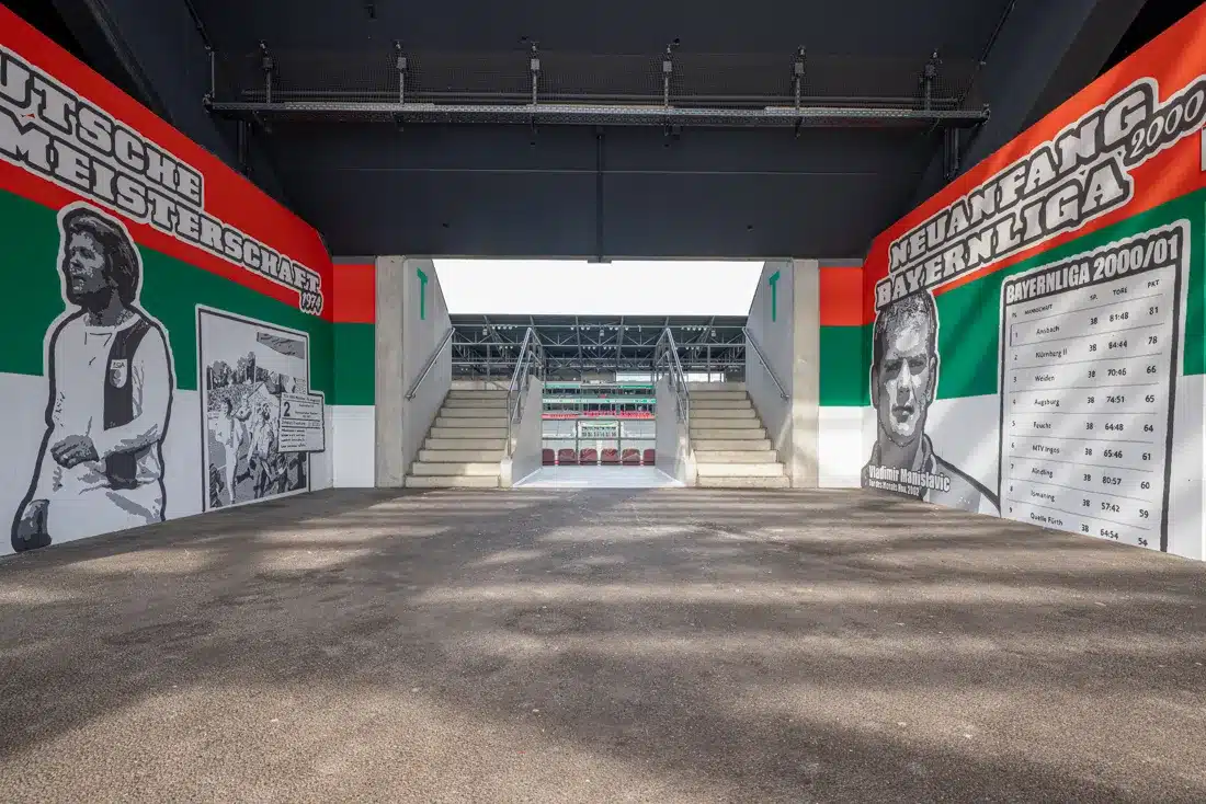 Zentraler Stadionzugang mit Wandmalereien: links "DEUTSCHE MEISTERSCHAFT 1974" und rechts "NEUANFANG BAYERNLIGA 2000" mit Tabelle und Porträts.